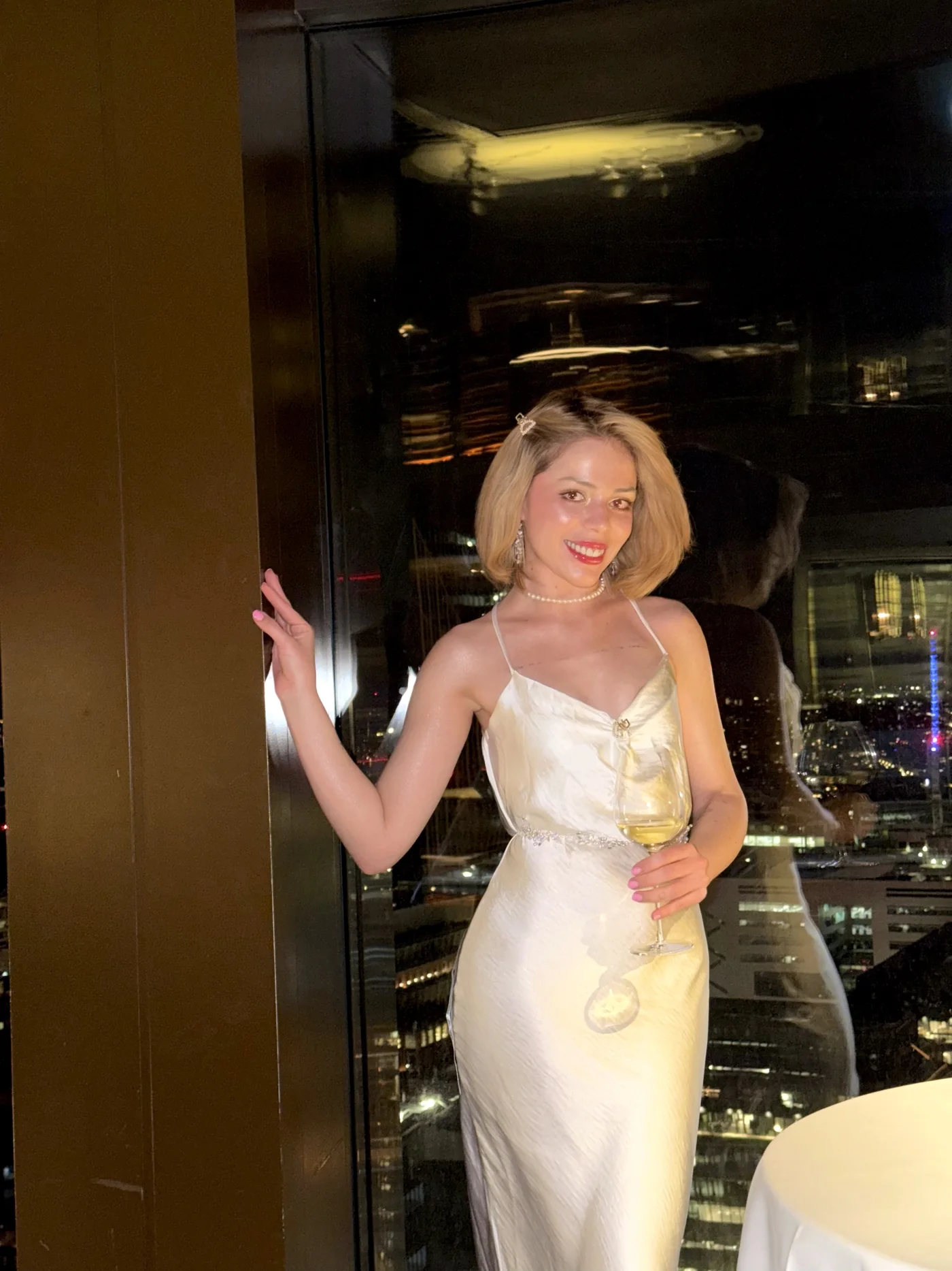 Laine en robe de satin blanc, un verre à la main, la ville derrière elle
